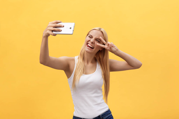 黄色の背景に分離された指で岩場の平和のシンボルを身振りで示すフロント カメラで selfie を撮影魅力的な明るい女の子のセルフ ポートレート - 写真・画像