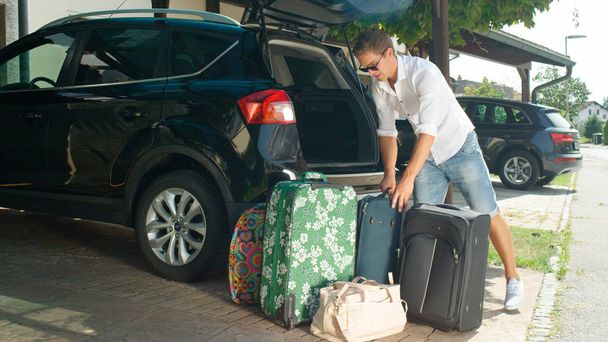 FLARE DE LENTES: Turista masculino descargando su equipaje del maletero de un gran coche negro estacionado frente a una casa suburbana. Hombre colocando bolsas de viaje en la acera de una calle soleada en un barrio tranquilo
. - Foto, Imagen