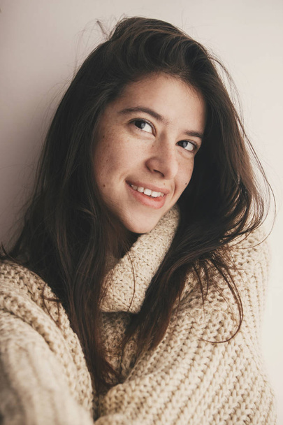Portrait of young attractive woman with no makeup and gorgeous hair in knitted sweater smiling in soft light in room. Stylish hipster girl taking selfie. Atmospheric cozy winter moments - Foto, immagini