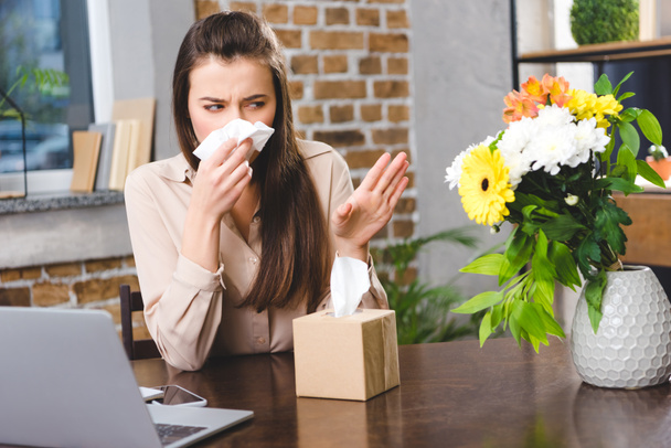 young businesswoman blowing nose and looking at flowers while suffering from allergy at workplace - Photo, Image