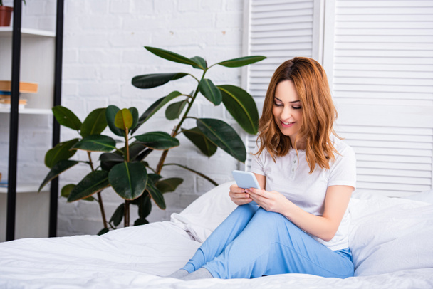 atraktivní žena s nazrzlé vlasy odpočívá v posteli a pomocí smartphone doma na víkend - Fotografie, Obrázek