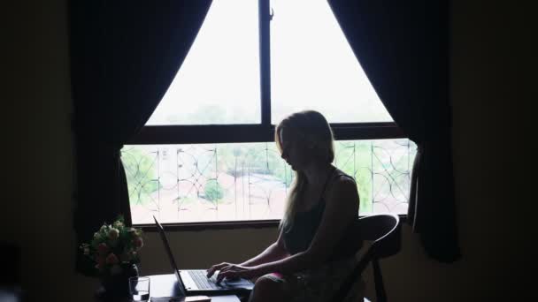 silhouette. girl sits on a chair by the window, uses her laptop. - Metraje, vídeo