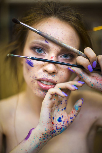 Εσωτερικη πορτρέτο του όμορφη γυναίκα καλλιτέχνης με βρώμικα χέρια με τα διαφορετικά χρώματα τους, κρατώντας πινέλα κοντά σε πρόσωπο και μάτια. - Φωτογραφία, εικόνα