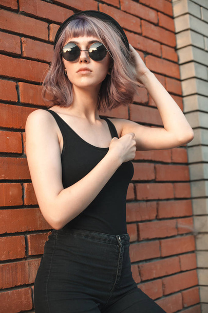 Young woman with hat and sunglasses is standing near the brick wall. - Foto, afbeelding