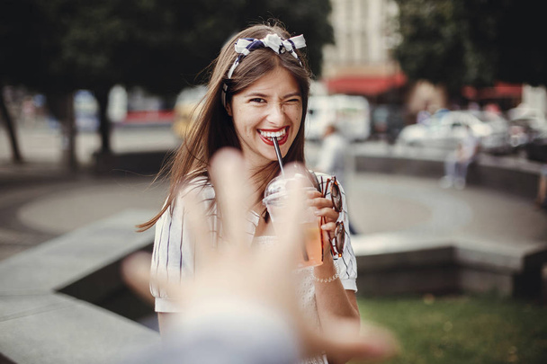 Hermosa mujer joven con estilo celebración de beber y relajarse en la calle de la ciudad. Chica hipster feliz en vestido retro y diadema, sosteniendo taza de plástico y paja, beber limonada
. - Foto, Imagen