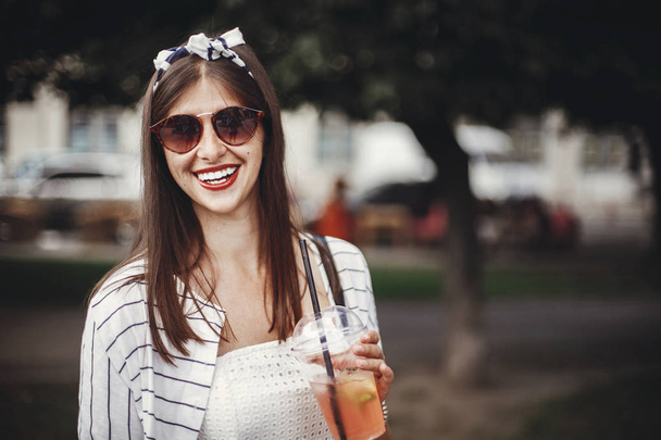 Bella elegante giovane donna con bevande fredde e sorridente in strada della città. Felice hipster ragazza con occhiali da sole, in abito retrò, tenendo monouso tazza di plastica e paglia
 - Foto, immagini
