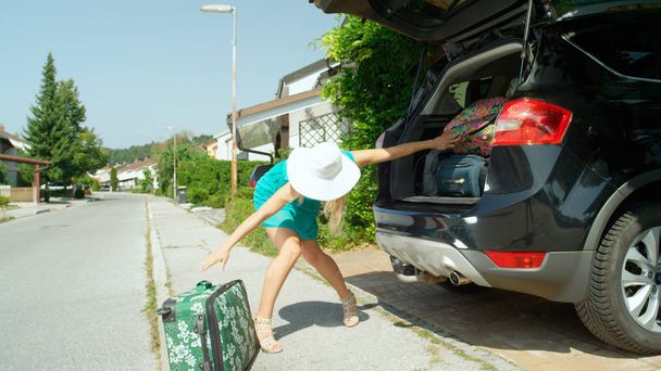 Jovem loira de salto alto carregando seu SUV preto com inúmeras malas antes de partir para suas férias de verão. Menina em turquesa sundress recheando o tronco de seu SUV para viagem de carro
. - Foto, Imagem