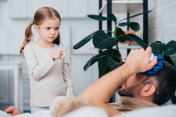 adorable daughter pointing on sick father touching head with ice pack in bedroom - Zdjęcie, obraz