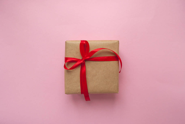 Gift box wrapped in brown colored craft paper and tied with red bow on pink background. - Photo, Image