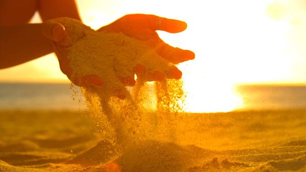 Nahaufnahme, Linsenschlag: Unerkennbare Touristinnen spielen mit dem warmen Sand bei Sonnenuntergang. malerische Nahaufnahme einer Sommerbrise, die der jungen Frau an einem goldenen Abend Sand aus den sanften Händen weht. - Foto, Bild