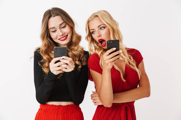 Portrait of two excited young smartly dressed women wearing makeup standing isolated over white background, looking at each other mobile phones - Фото, изображение