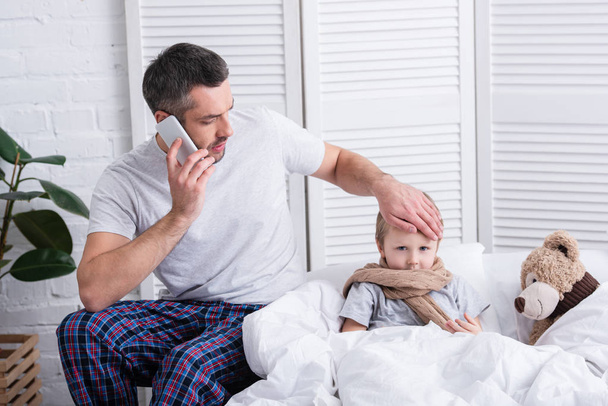 padre che parla con smartphone e tocca la fronte del figlio malato in camera da letto
 - Foto, immagini