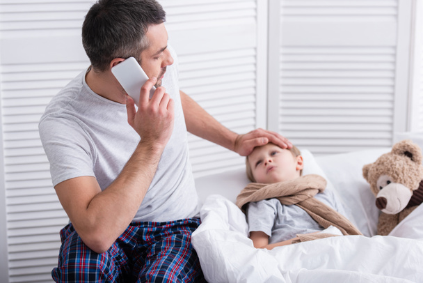 papà che parla con smartphone e tocca la fronte del figlio malato in camera da letto
 - Foto, immagini