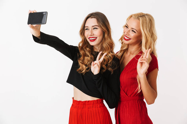 Portrait of two cheerful young smartly dressed women wearing makeup standing isolated over white background, taking a selfie - Photo, image