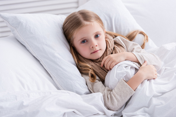 niño enfermo con bufanda sobre el cuello acostado en la cama en casa y mirando a la cámara
 - Foto, Imagen