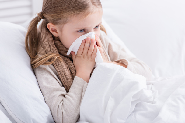 sick child with scarf over neck lying in bed and blowing nose in tissue at home - Zdjęcie, obraz