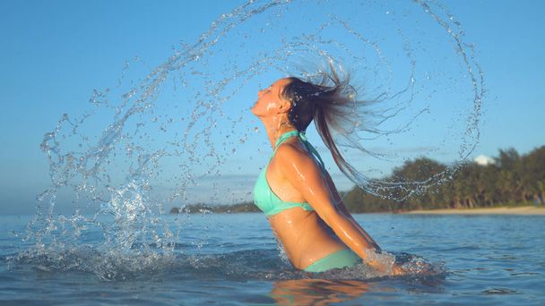 CLOSE UP: Playful Caucasian woman in turquoise bikini splashes glassy ocean water by flipping her hair back. Girl on summer vacation plays in the refreshing sea close to beautiful tropical island. - Foto, Bild
