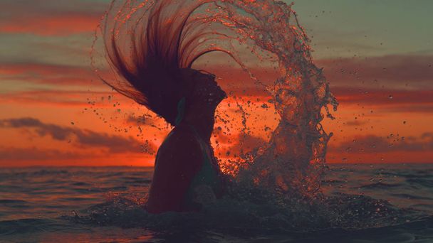CERRAR, SILHOUETTE: Chica irreconocible voltea su cabello hacia atrás y rocía agua fresca del océano sobre la pintoresca puesta de sol naranja. Desconocida turista femenina relajándose en lo exótico en la noche espectacular
. - Foto, imagen