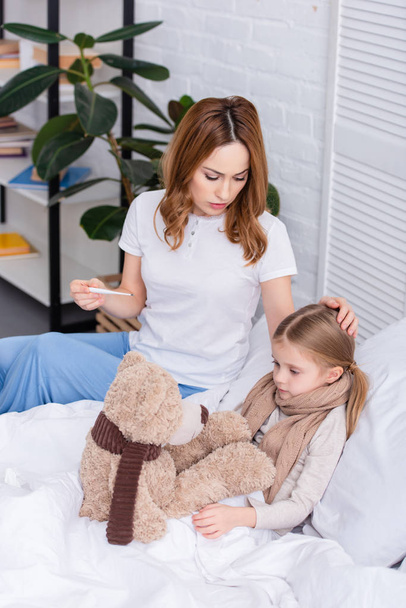 mommy taking care of sick daughter in bedroom and checking her temperature with thermometer - Foto, Imagem