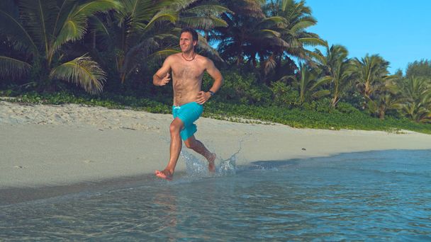 Активный человек на удивительных летних каникулах бегает вдоль идиллического белого песчаного берега. Молодой белый мужчина наслаждается расслабляющей пробежкой по солнечному тропическому пляжу. Пригодный турист брызгает стеклянную воду
 - Фото, изображение