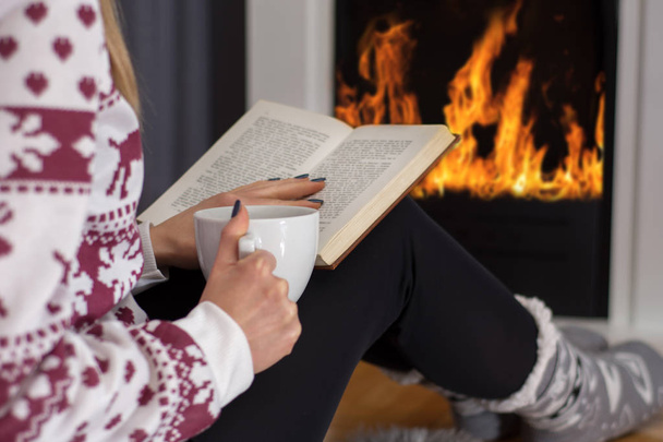 Młoda dziewczyna, siedząc przy kominku, czytając książkę i picie gorącej herbaty. Dziewczyna ocieplenia nóg na ogień i nosi Skarpety wełniane i sweter. Zima i zimno. Zbliżenie, Selektywny fokus - Zdjęcie, obraz