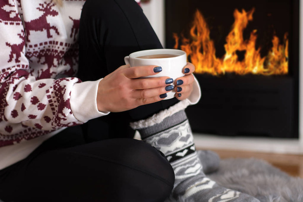 Niña sentada frente a la chimenea y sosteniendo té de bebida caliente en la mano en casa en la larga noche de invierno. La mujer usa suéter de lana y calcetines. Concepto de invierno y clima frío. Primer plano, enfoque selectivo
 - Foto, imagen