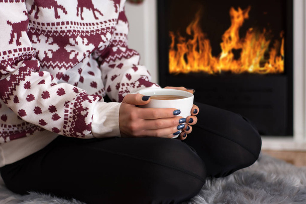 Dziewczyna, picie gorącej herbaty przy kominku i siedzi na furry dywanów w domu. Kobieta nosi wełniany sweter. Zima i zimno koncepcja. Zbliżenie, Selektywny fokus - Zdjęcie, obraz