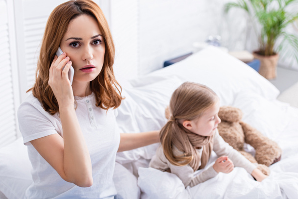 scared mother calling doctor by smartphone, sick daughter in bed - Photo, Image