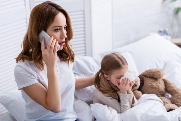 Matko volající lékaře o smartphone, nemocná dcera foukání nosem v posteli - Fotografie, Obrázek