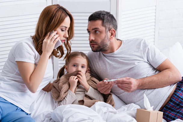 mom calling doctor and dad checking temperature of sick daughter in bedroom - Photo, image