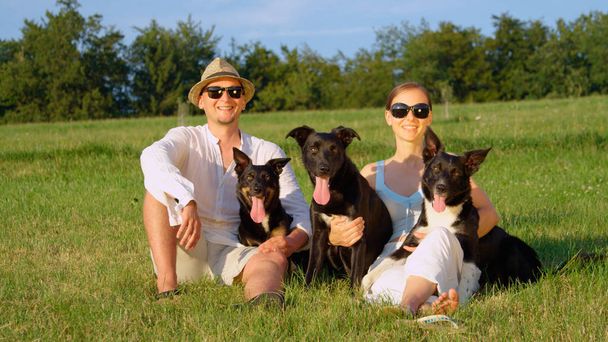PORTRAIT: Happy couple poses sitting in the empty grassy field with their three adorable border collie dogs on a sunny day in summer. Cheerful young man and woman enjoying the outdoors with puppies. - Photo, Image