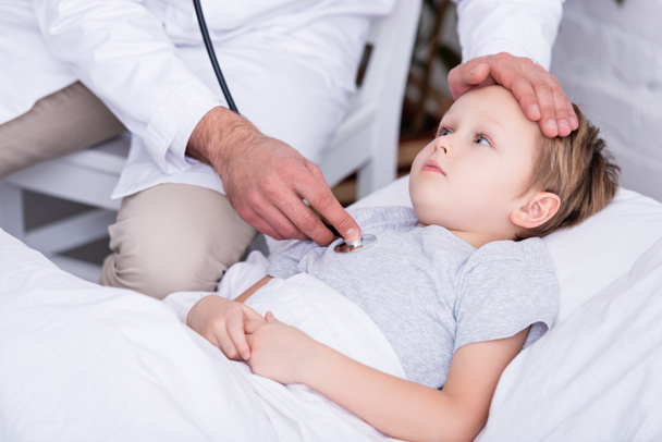 imagen recortada de pediatra de capa blanca examinando niño enfermo con estetoscopio y tocando la frente
 - Foto, Imagen