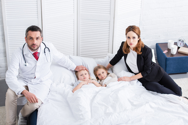 pediatrist and mother sitting on bed with two sick kids and looking at camera in bedroom - Photo, Image