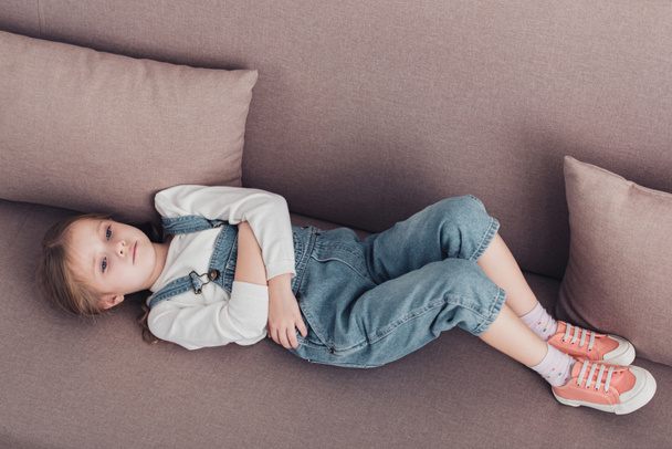 высокий угол обзора больного ребенка с болью в животе лежит на диване в гостиной
 - Фото, изображение