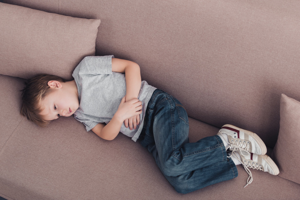 high angle view of sick boy with stomach pain lying on sofa in living room - Φωτογραφία, εικόνα