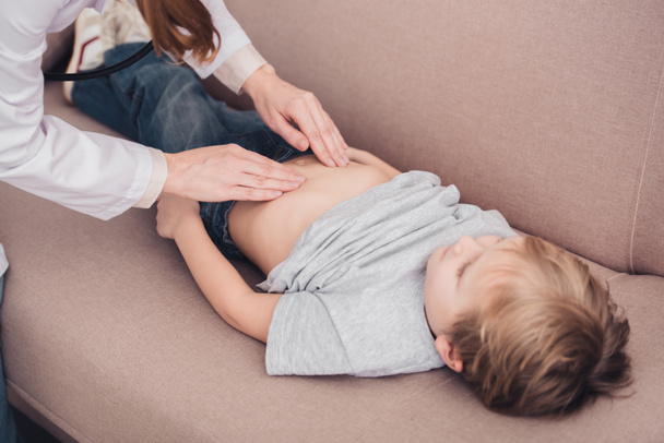 cropped image of pediatrist in white coat palpating sick boy stomach on sofa in living room - Φωτογραφία, εικόνα