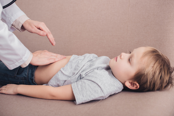 levágott kép tapogató beteg fiú gyomor a nappaliban kanapéval pediatrist - Fotó, kép