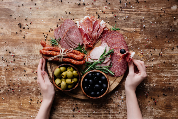 cropped view of  woman taking prosciutto from round cutting board with olives, salami and ham on wooden table with scattered peppercorns - Фото, зображення