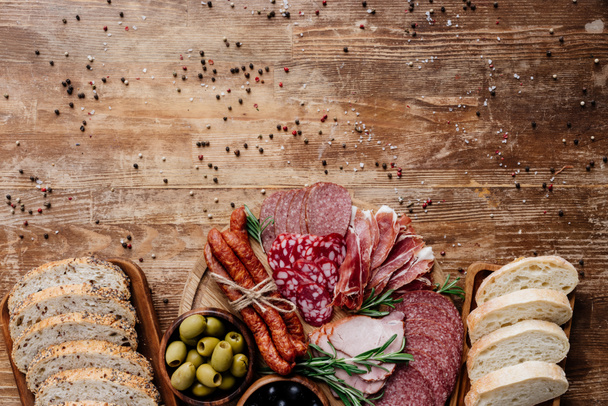 top view of cutting boards with delicious bread, salami, smoked sausages, ham and olives on wooden table with scattered peppercorns - Foto, Imagem