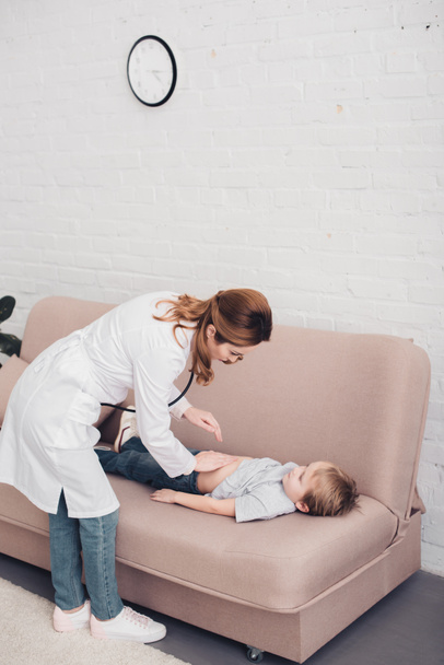pediatrist tapogató beteg fiú gyomor a nappaliban kanapé magas szög kilátás - Fotó, kép