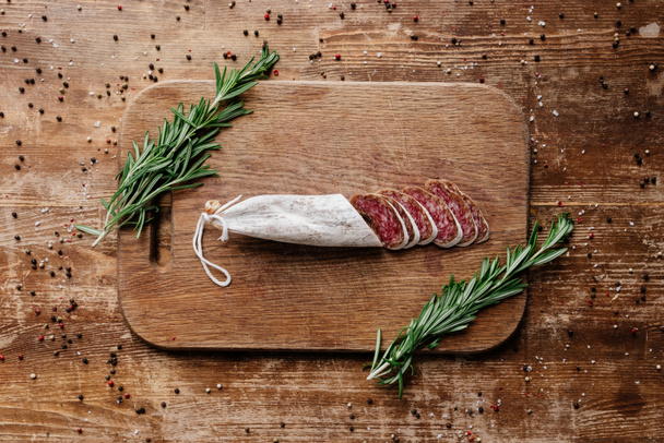 top view of cutting board with rosemary and delicious sliced salami on wooden table with scattered spices - Foto, Imagem