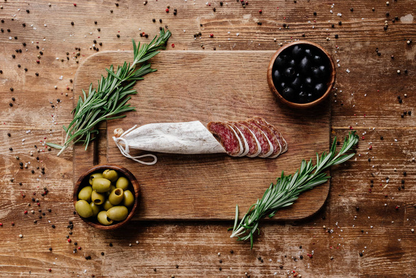 top view of cutting board with rosemary, black and green olives in two bowls and delicious sliced salami on wooden table with scattered spices - Valokuva, kuva