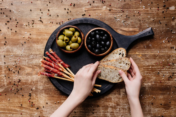 cropped view of  woman putting bread on round cutting board with olives in two bowls and breadsticks with prosciutto on wooden table - Photo, image