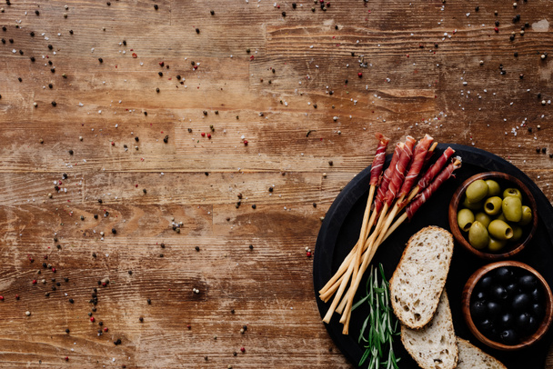 κάτοψη του μαύρο μαρμάρινο κοπής με ελιές σε μπολ, κριτσίνια, προσούτο, ψωμί και μυρωδικά σε ξύλινο τραπέζι με διάσπαρτα κόκκους πιπεριού - Φωτογραφία, εικόνα