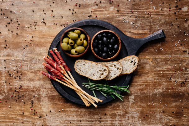 top view of black marble cutting board with olives, breadsticks, prosciutto, bread and rosemary on wooden table with scattered peppercorns - Photo, Image