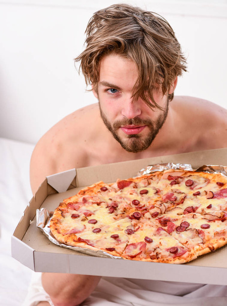 Portrait of lazy muscular man eating pizza while laying on a bed at home. Man bearded handsome bachelor eating cheesy food for breakfast in bed. Who cares about diet. Eating pizza in bed. - Foto, Imagem