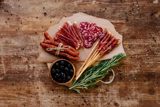 top view of round cutting board with black olives, delicious prosciutto, salami, smoked sausages and herbs on wooden vintage table with scattered peppercorns - Фото, изображение