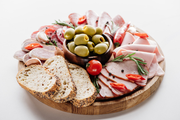 round cutting board with delicious salami, ham, olives in bowl, bread, herbs and vegetables on white table - Photo, Image