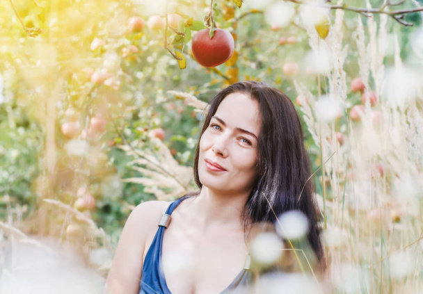 femme dans une robe d'été légère est assis dans l'herbe sèche dans le verger de pommes en regardant une pomme rouge sur une branche. Séduction, sexualité. Santé des femmes. Reproduction. Naissance des enfants
. - Photo, image
