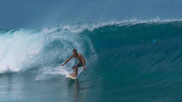 アクティブな若い男のサーフィンが彼の楽しみの中に夏の完璧な日に大きなターコイズ バレル ウェーブ休暇の楽園の島の近く。フランス領ポリネシアのスポーツマン サーフィン壮大な海の波の息をのむショット. - 写真・画像